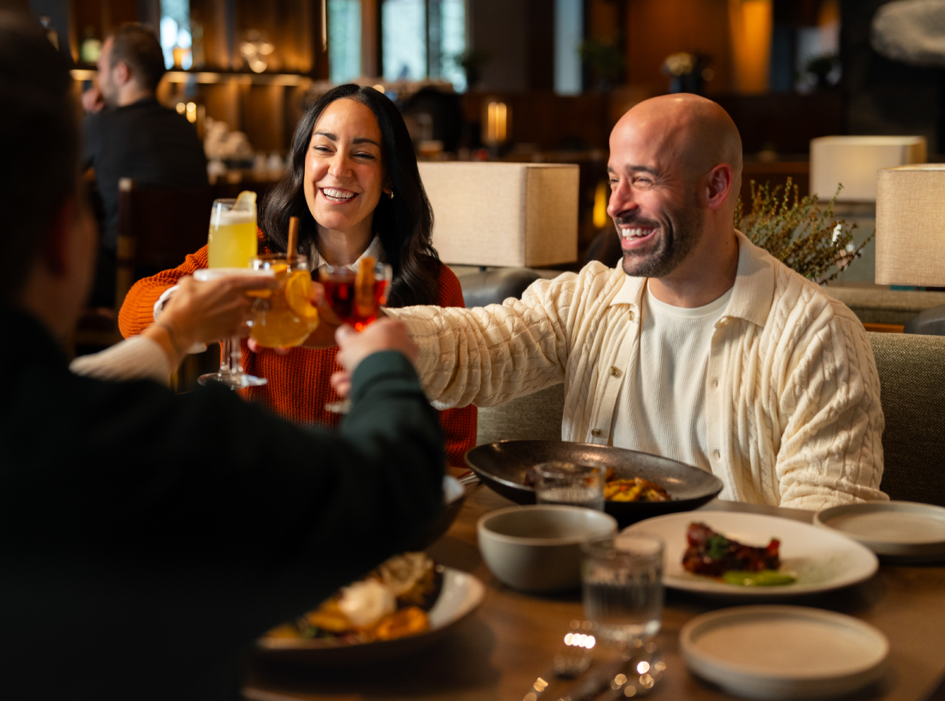 A group cheers with cocktails at the Good Folk restaurant, smiling and laughing. warm, inviting atmosphere, upscale decor, evening lighting