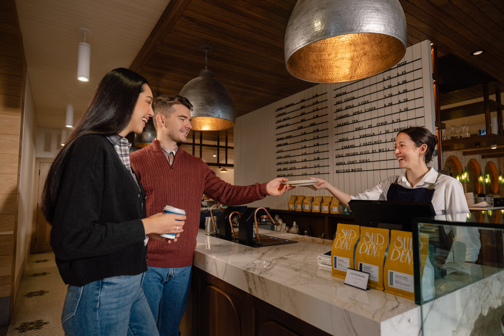 a couple holding coffee cups from sudden sally interacting with barista at the till area. warm ambient lighting, coffee menu board, and bagged coffee at the counter